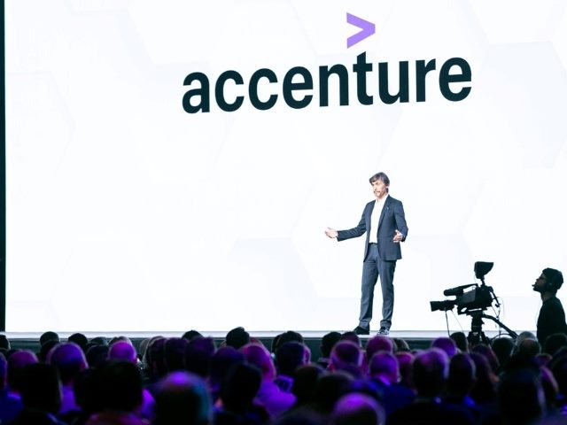 Sitecore CEO Steve Tzikakis on stage with an accenture logo on the screen behind him at his company's 2022 Sitecore Symposium conference.