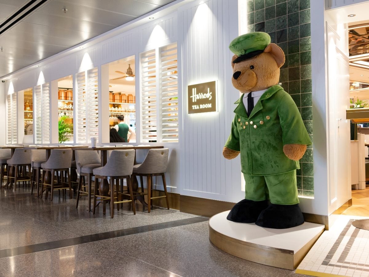 Large teddy bear statue dressed in a green uniform stands outside the Harrods Tea Room, with bar seating and a bright, upscale interior visible behind it.