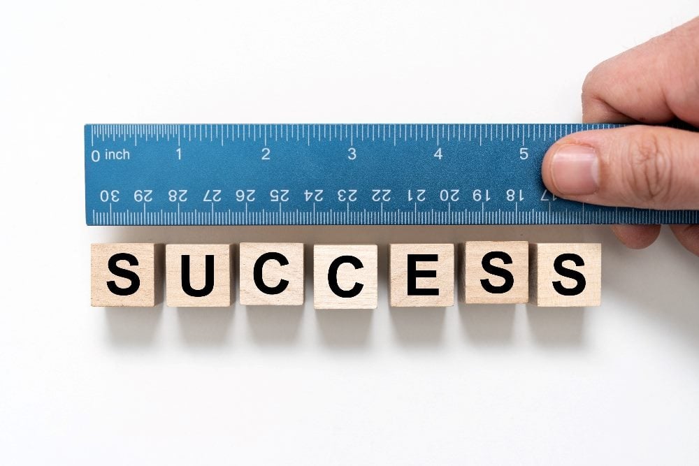 A hand holds a blue ruler with white numbers with wooden blocks below that spell out the word "success" in piece about the need for change in customer experience measurement and employee satisfaction factors among others.