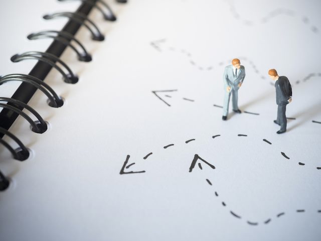 miniature figures of two businessmen in suits standing on a notebook with arrows around them