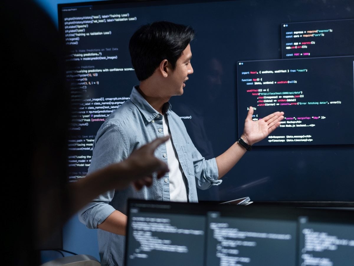 A software developer presents code displayed on large digital screens during a team meeting. Multiple monitors show programming scripts and application code as colleagues collaborate in a modern technology workspace.