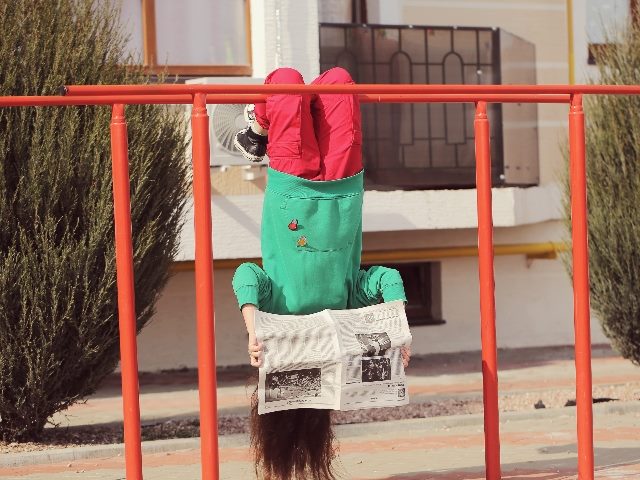 A girl in bright clothes reads a newspaper on the street while hanging upside down on a playground apparatus. 