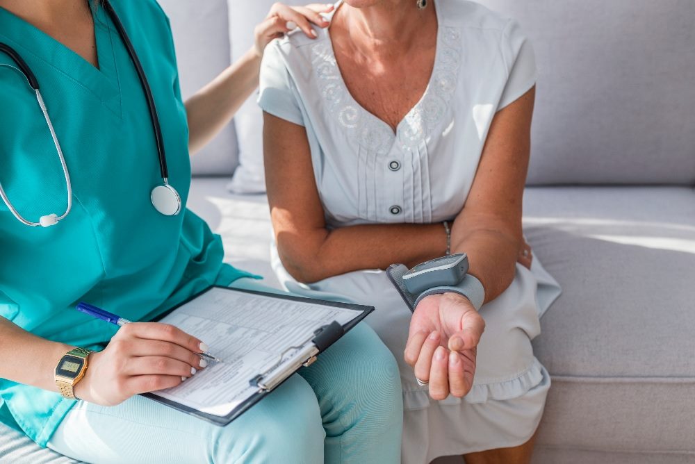 A nurse sits on a couch with a patient and puts hand on the patient's shoulder while holding a clipboard and taking their blood pressure in story about peak-end rule and better customer experiences.