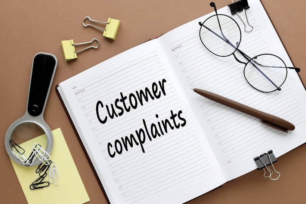 The words customer complaints appears on an open diary with a pen and wire rim glasses visible as well as paper clips and sticky notes in piece about CX audits.