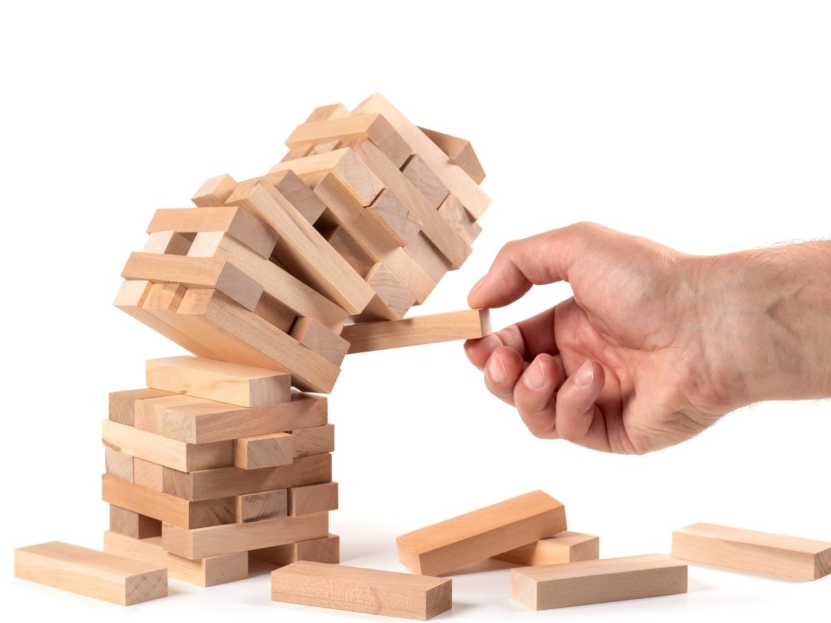 A hand pulls a wooden block from the base of a Jenga-style tower, causing the upper section of stacked blocks to tilt and topple sideways as several loose blocks lie scattered on the white surface below.