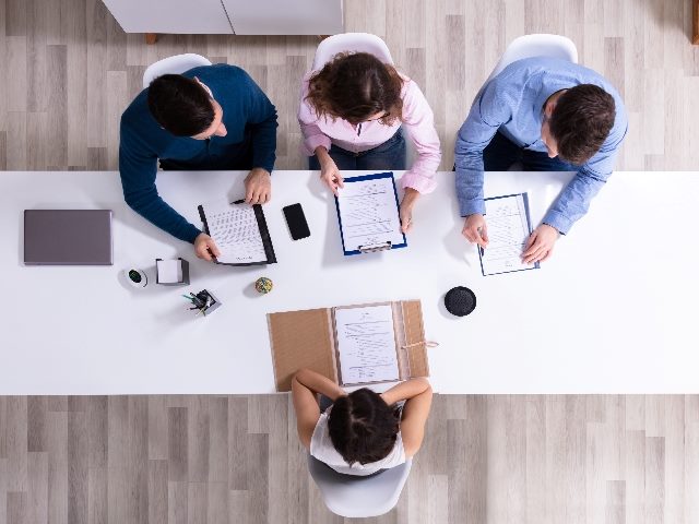 Aerial view of a job interview, with one woman sitting on one side of the desk and three interviewers sitting on the other side.