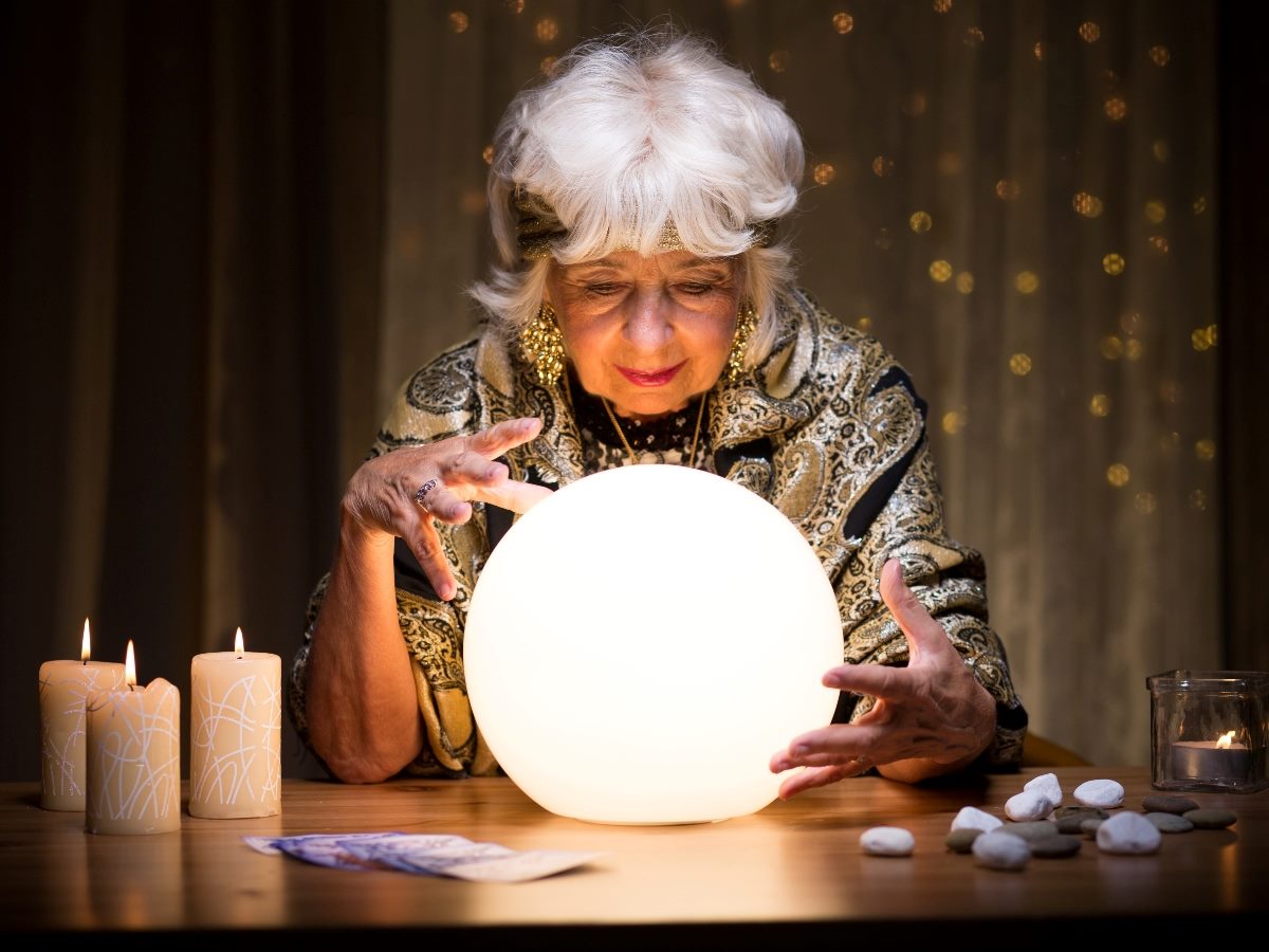 An older woman dressed in ornate, patterned clothing leans over a glowing crystal ball, holding her hands around it as lit candles and small stones sit on the table in front of her.