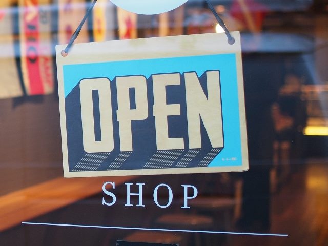 An na open sign on a turquoise background with white letters hangs on a glass door to a shop with the word "shop" below the sign in piece about the GPT Store launch. 