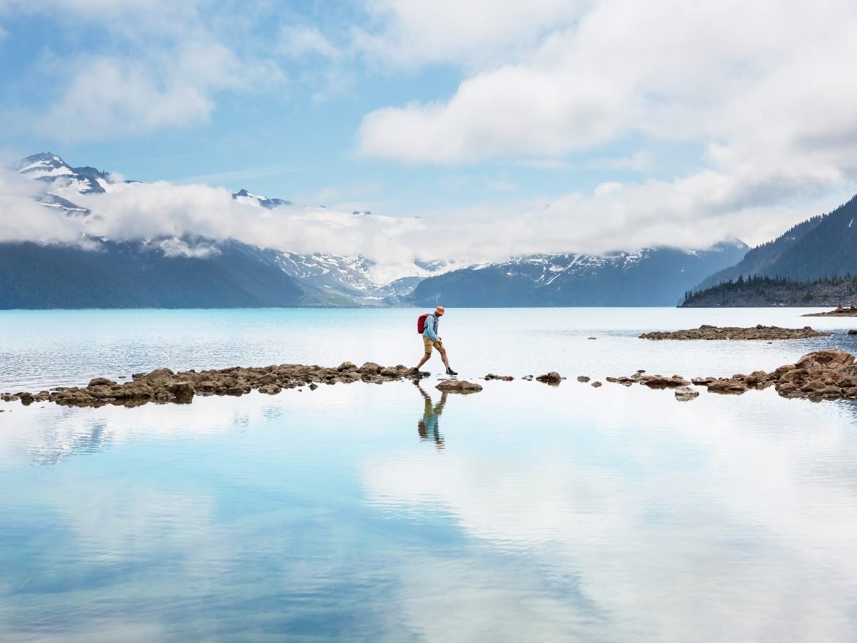 A hiker with a red backpack crosses a rocky path over calm blue water, surrounded by snow-capped mountains and low clouds reflecting in the lake.