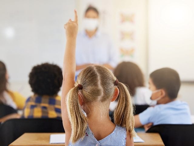 Young girl wearing a blue dress and with her hair in pony tails sits at a desk in a school classroom and holds up her hand with her teacher in the background in piece about well-crafted FAQs.