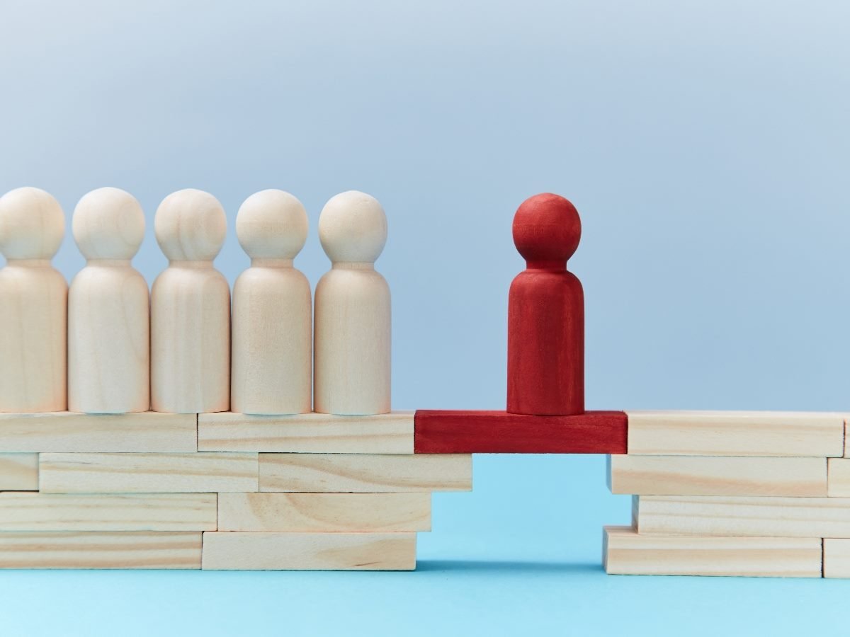 A red wooden figure stands on a red bridge connecting two stacks of wooden blocks, linking a line of neutral-colored figures to the other side — symbolizing leadership, connection and bridging gaps in decision-making.