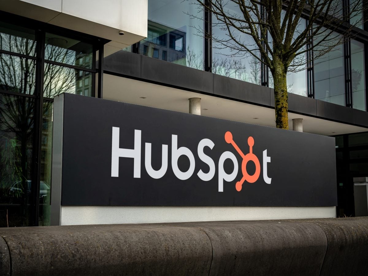 A dark gray monument sign reading "HubSpot" in white lettering with the brand's orange node-and-line logo replacing the "o," mounted on a low concrete wall outside a glass-and-steel office building, with bare tree branches and reflected windows visible in the background.