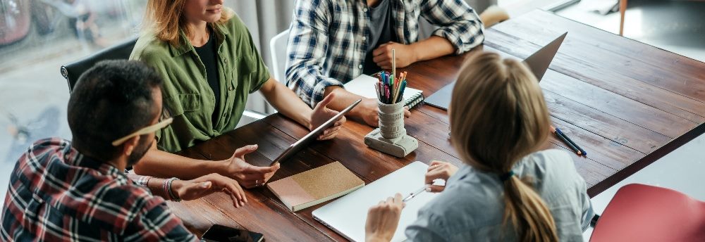 Workers sit at a table with papers and laptop and collaborate on a project suggesting the importance of customer experience models.