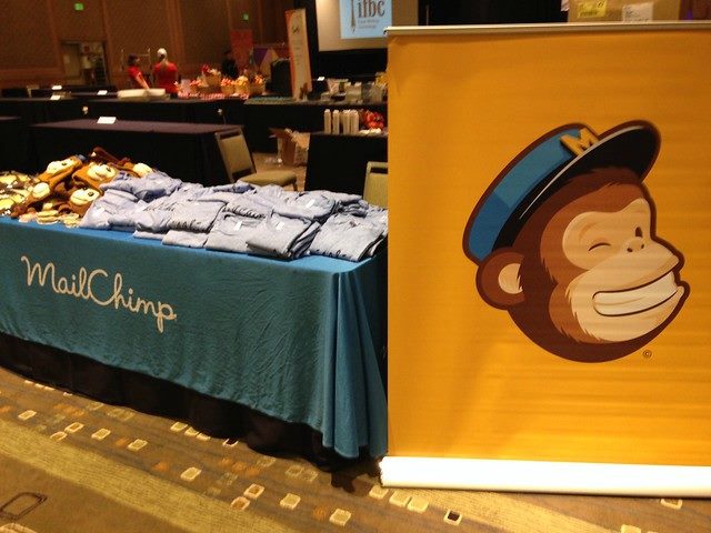 A table with a Mailchimp tablecloth and Mailchimp-theme shirts on top, next to a post with the Mailchimp logo on it. 