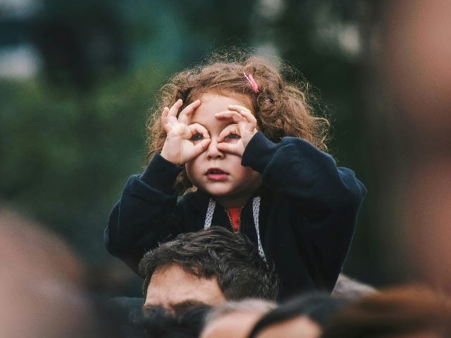 Girl sitting on someone's shoulders using hands to pretend she's using binoculars.