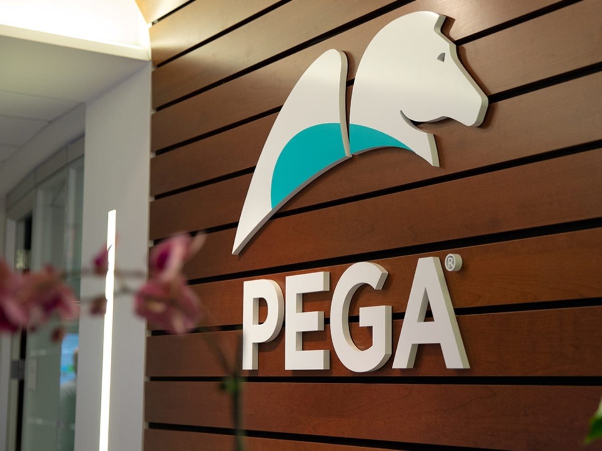 Large PEGA logo mounted on a wood-paneled office wall, featuring the company's white horse-head emblem with a teal accent above bold white lettering. Blurred plants frame the foreground of the modern corporate workspace.