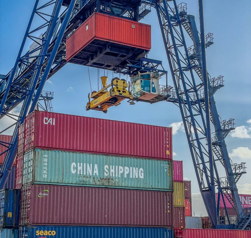 Shipping containers stacked, one with "China shipping" written on it, as they await a large crane device to lower another container on top of the stack.