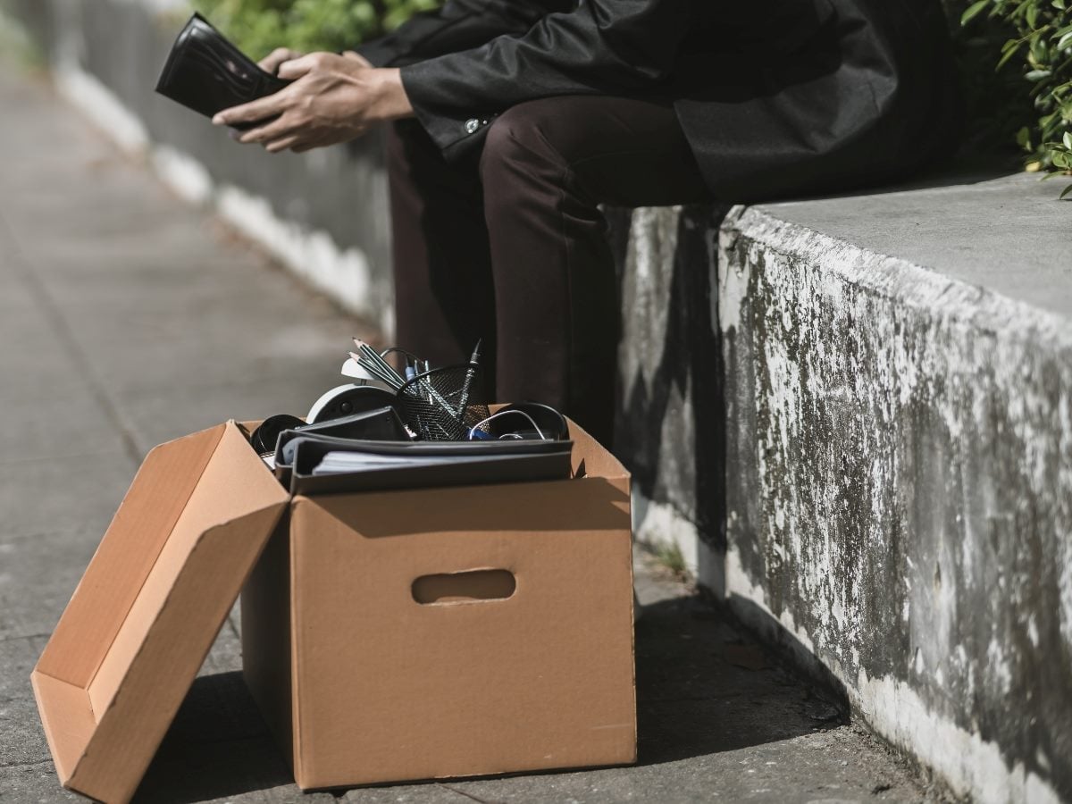 A person in business attire sits on a concrete ledge holding a wallet, with a cardboard box of office items placed beside them on the sidewalk.