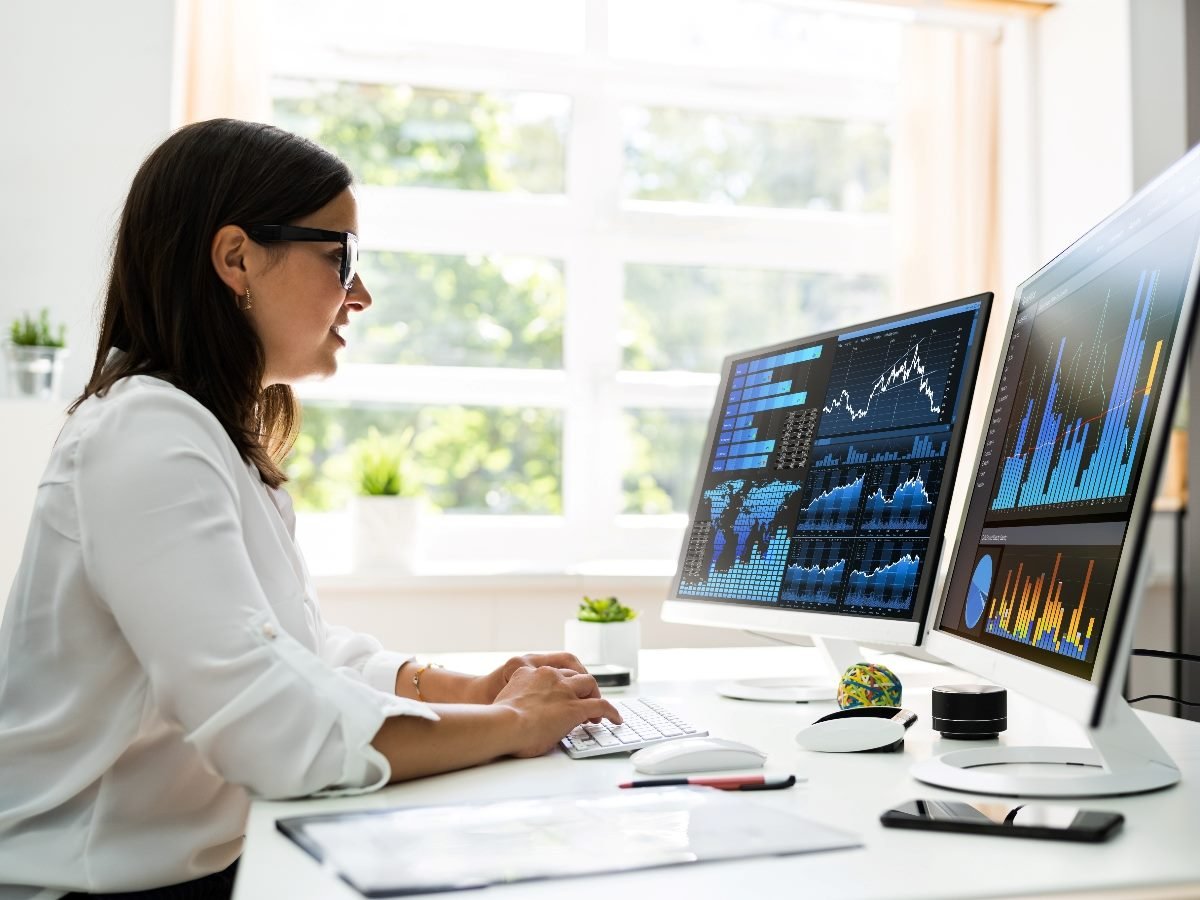 A business professional analyzes data on dual computer monitors displaying colorful charts, graphs and analytics dashboards in a bright modern office.