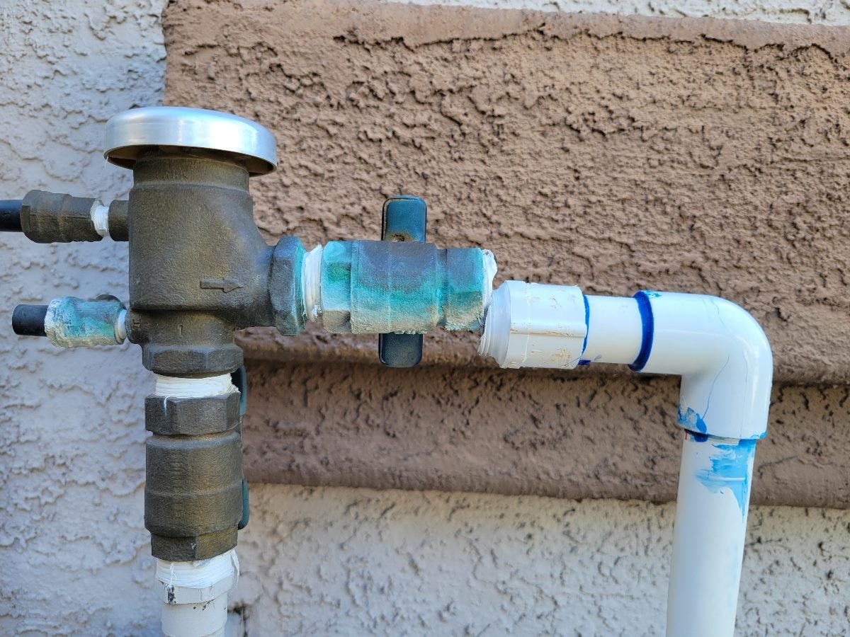 Close-up of an outdoor plumbing connection showing a corroded brass backflow preventer attached to white PVC pipes, with visible blue-green oxidation and sealant residue around the joints.