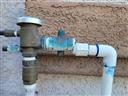 Close-up of an outdoor plumbing connection showing a corroded brass backflow preventer attached to white PVC pipes, with visible blue-green oxidation and sealant residue around the joints.