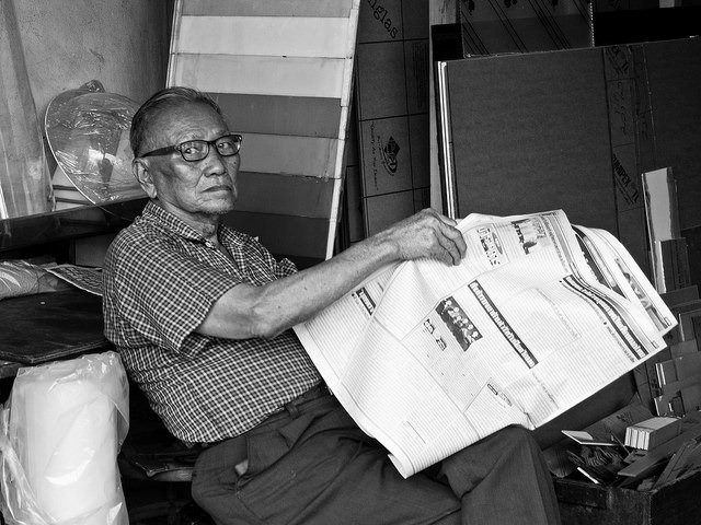 Man sitting down looking at the camera as he turns the page in a newspaper.