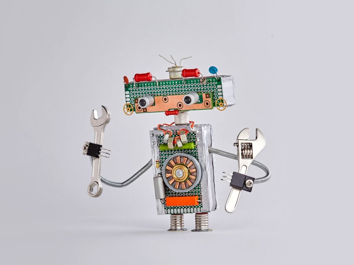 A whimsical robot assembled from electronic components, circuit boards, wires and metal hardware stands against a light gray background. The handmade robot has googly eyes, a circuit-board head and body, spring-like arms and legs, and holds two wrenches as if ready to perform repairs or maintenance.
