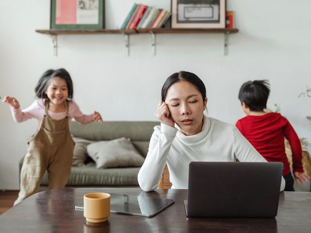 exhausted woman on laptop with two very active kids behind her