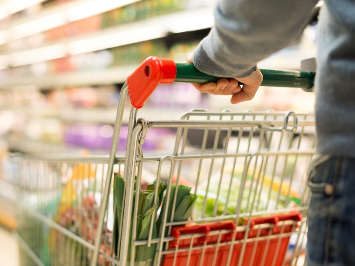 Close-up of a person’s hand pushing a grocery shopping cart down a supermarket aisle, with shelves of products blurred in the background.