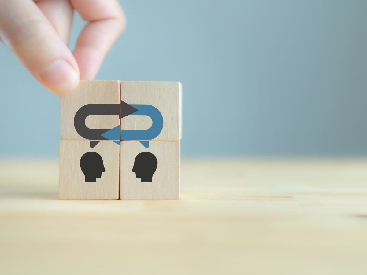 A hand places a wooden block onto a stack of cubes featuring two silhouetted heads and a circular arrow symbol, representing alignment, communication and the continuous exchange of information between teams or stakeholders.