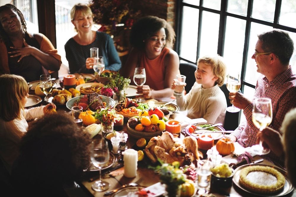 A group of adults and children sit around a dinner table filled with Thanksgiving-style food, laughing and raising glasses in a bright, cozy dining room.
