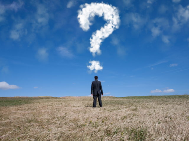 A businessman standing in an open filed with his back turned to the camera. Above his head there is a cloud shaped like a question mark in the sky. - big questions concept