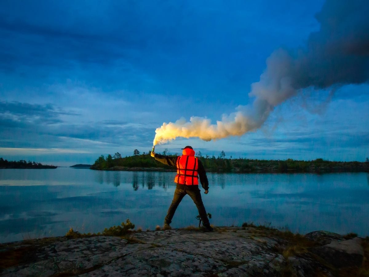 Person standing on a rocky shoreline holding a lit flare that releases a plume of smoke over a calm lake at dusk, symbolizing a signal or warning.