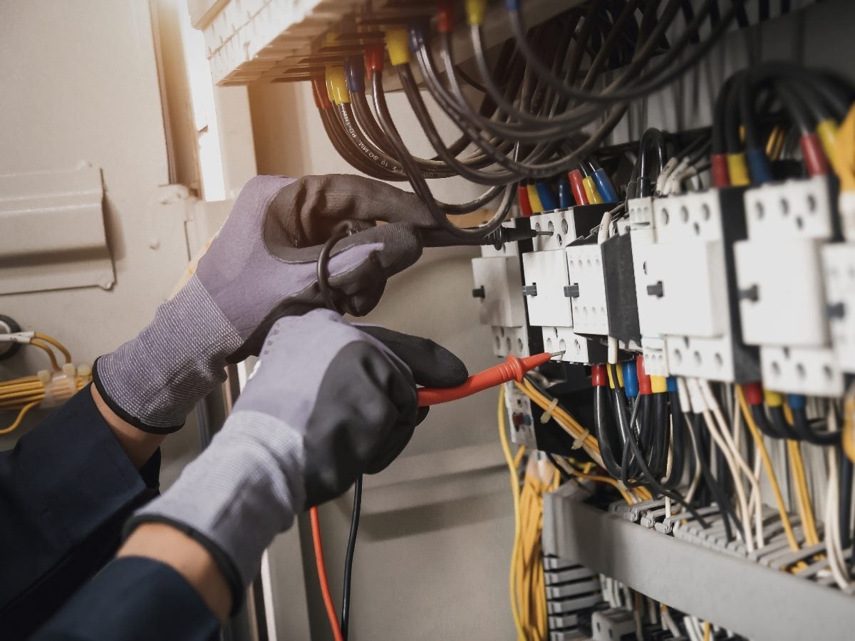 A technician wearing protective gloves uses electrical testing probes inside an industrial control panel filled with wiring, breakers and electrical components during maintenance or troubleshooting work.