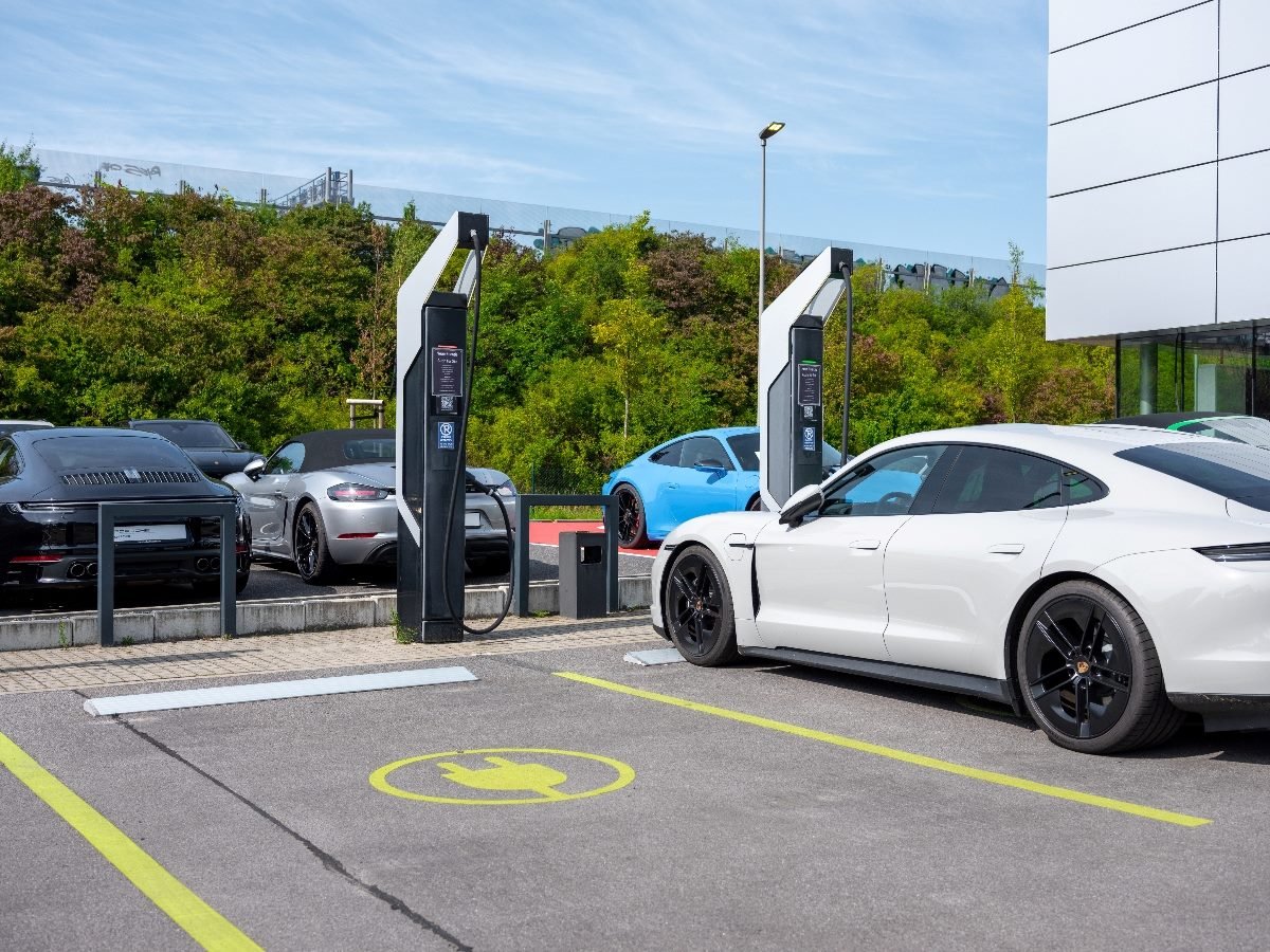 Electric cars charging at modern charging stations in a parking lot, including a white sedan in the foreground and multiple sports cars in the background.