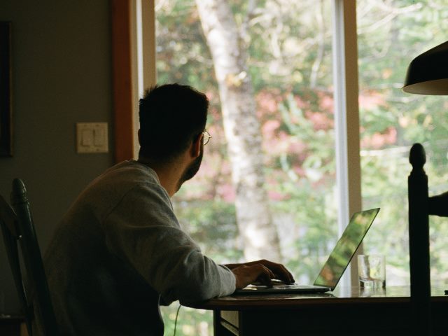 man in gray hoodie looking out a window while using a laptop at home