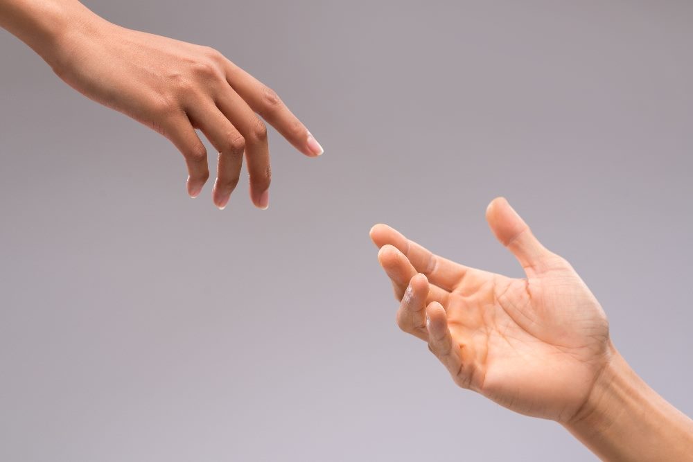 Two artistically lit hands reach out for each other from a top corner on the left and a bottom, corner from the right against a gray background in piece about the future of customer interactions.