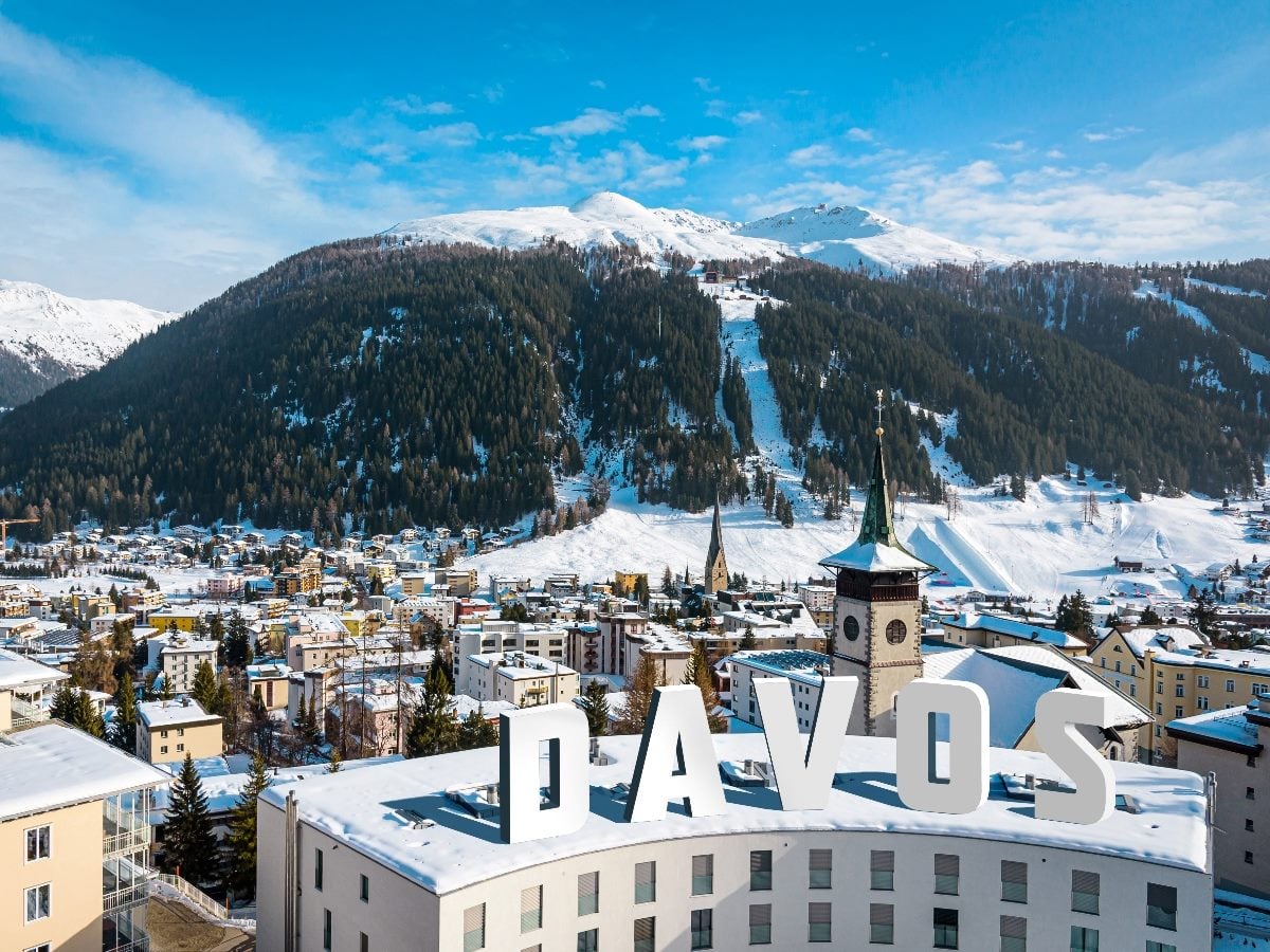 Snow-covered Davos, Switzerland, with alpine mountains rising behind the town center and a prominent “DAVOS” sign atop a building under a clear blue winter sky.