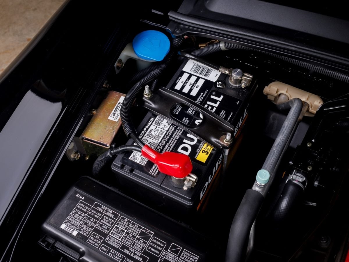 Close-up of a Duracell car battery installed in a vehicle engine bay, showing the battery terminals, wiring, and surrounding engine components.