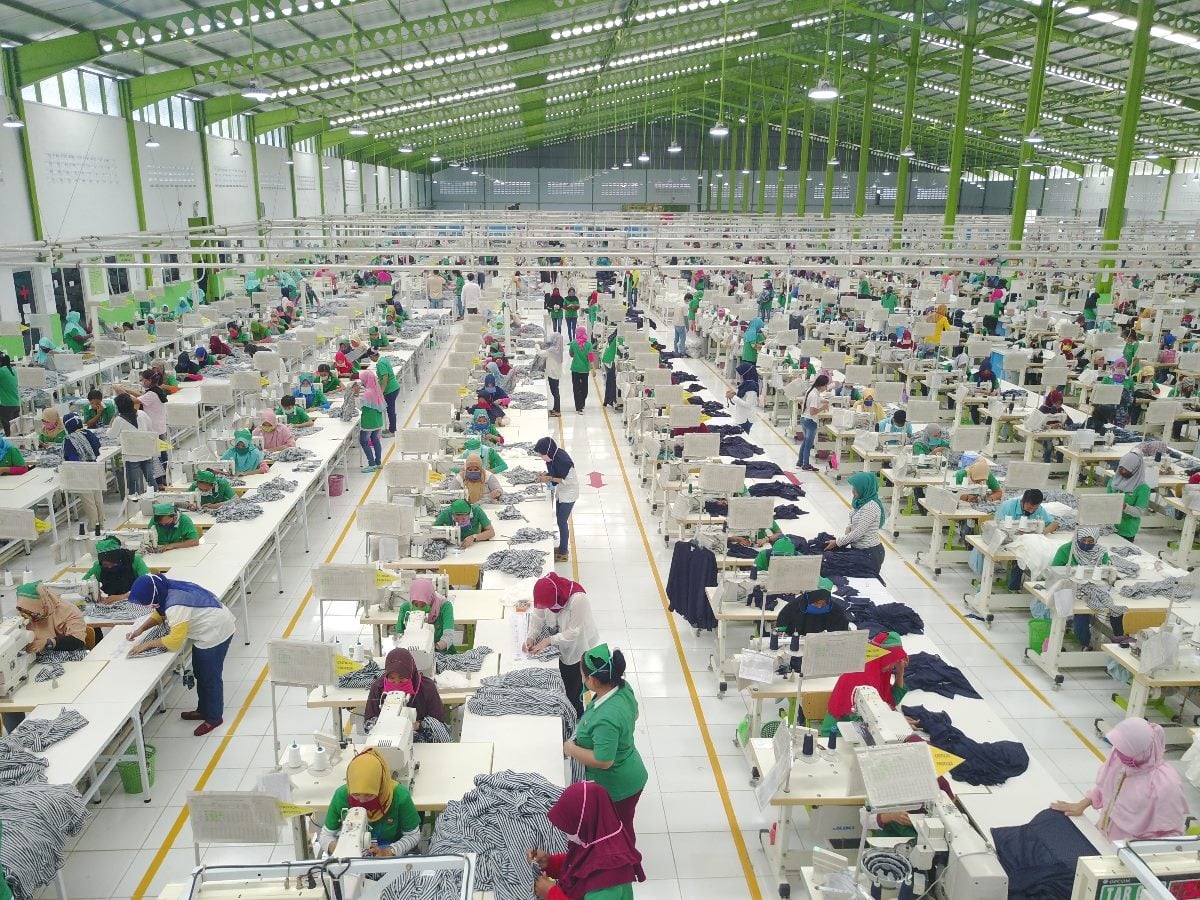 Workers in a large garment factory floor, rows of sewing machines stretching across a brightly lit industrial space with employees assembling clothing at scale.