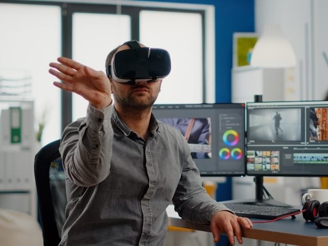 A man wearing a virtual reality headset sitting down and point to the right with his right hand. 