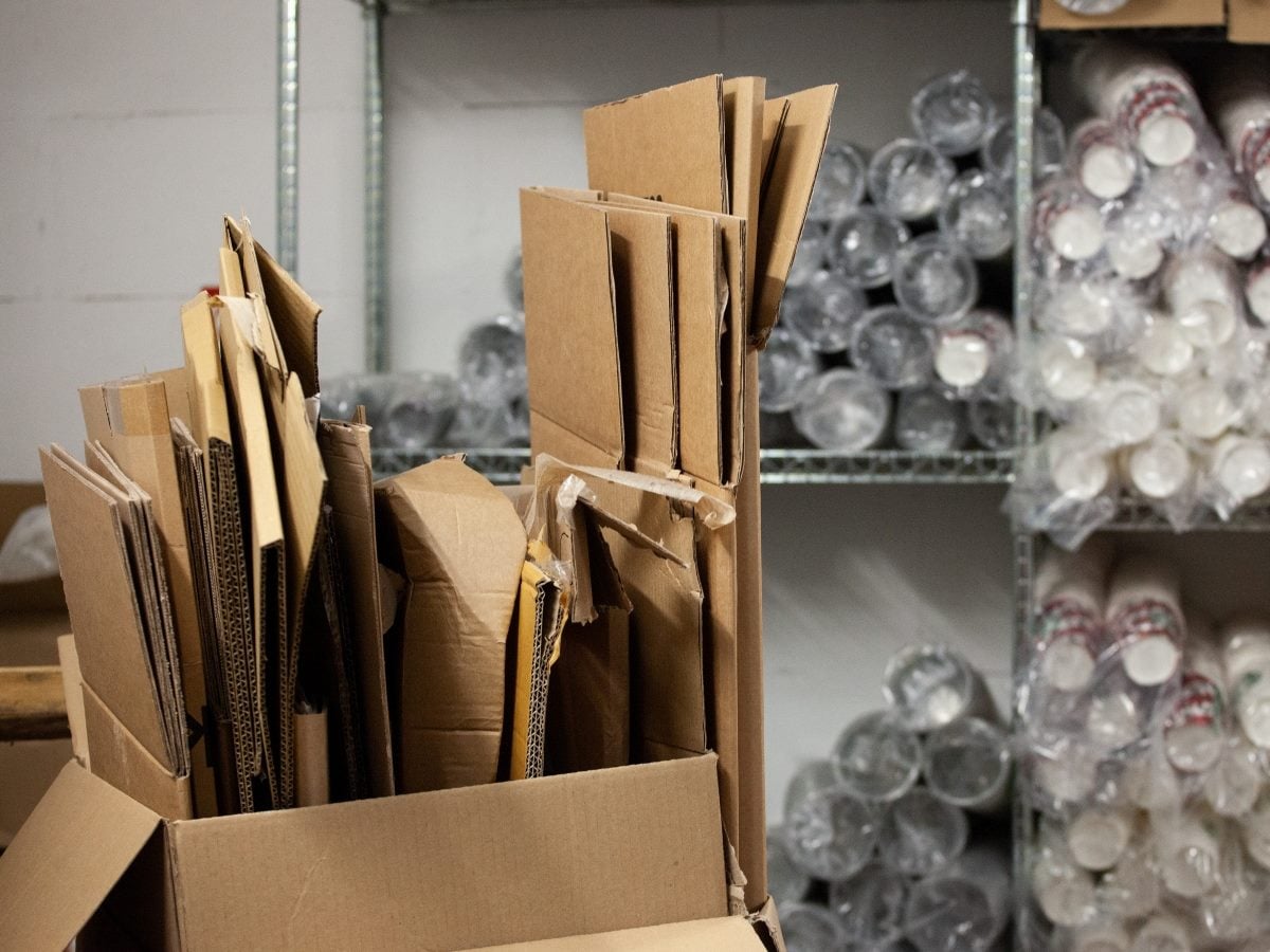 Flattened cardboard boxes stand upright in a large open box inside a warehouse or storage area, with shelves of stacked clear plastic cups wrapped in plastic visible in the background. The image highlights recycled packaging materials awaiting reuse or disposal.