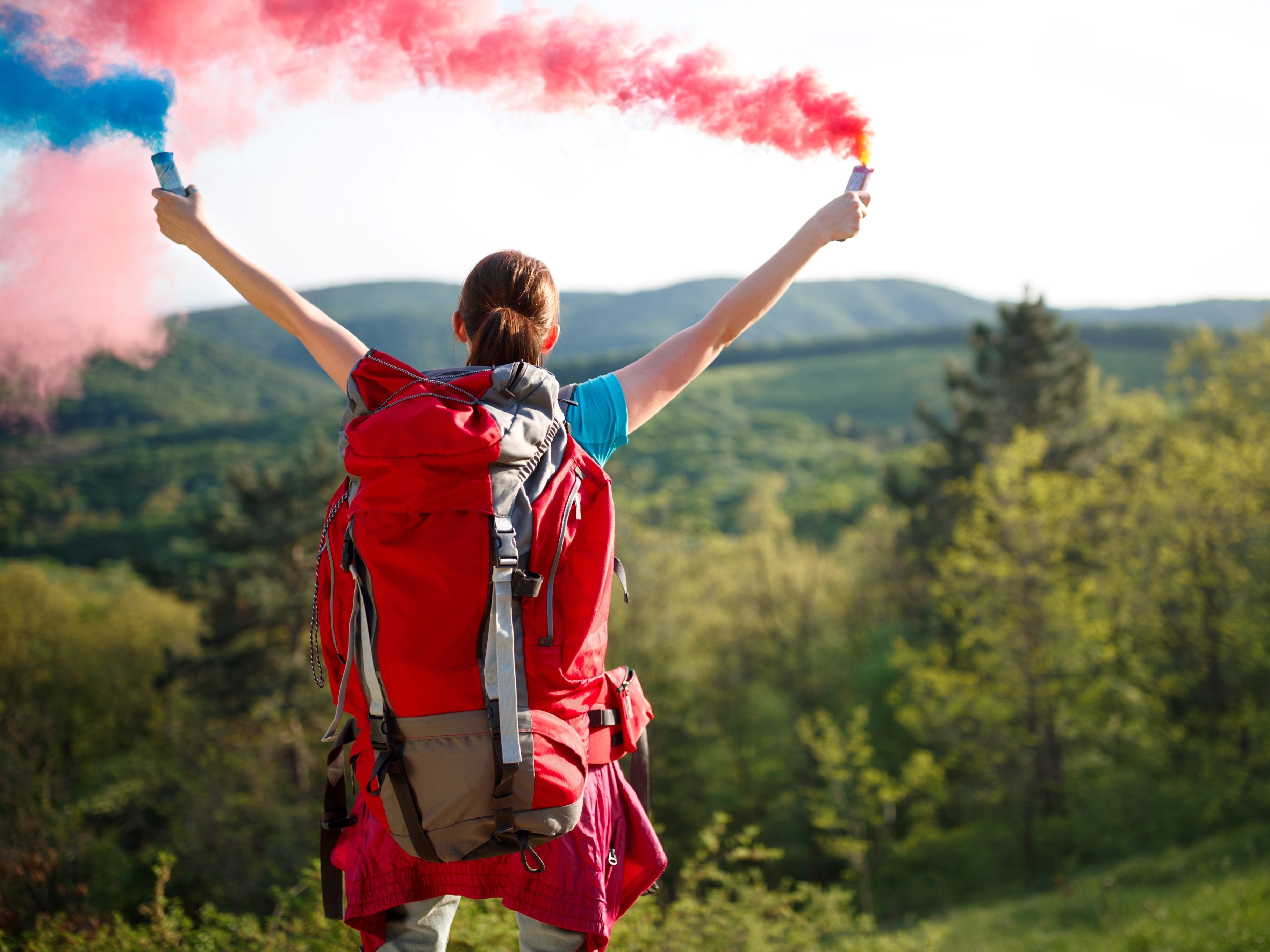 Hiker wearing a red backpack stands on a hilltop overlooking a forested landscape, holding blue and red smoke flares aloft against a clear sky.