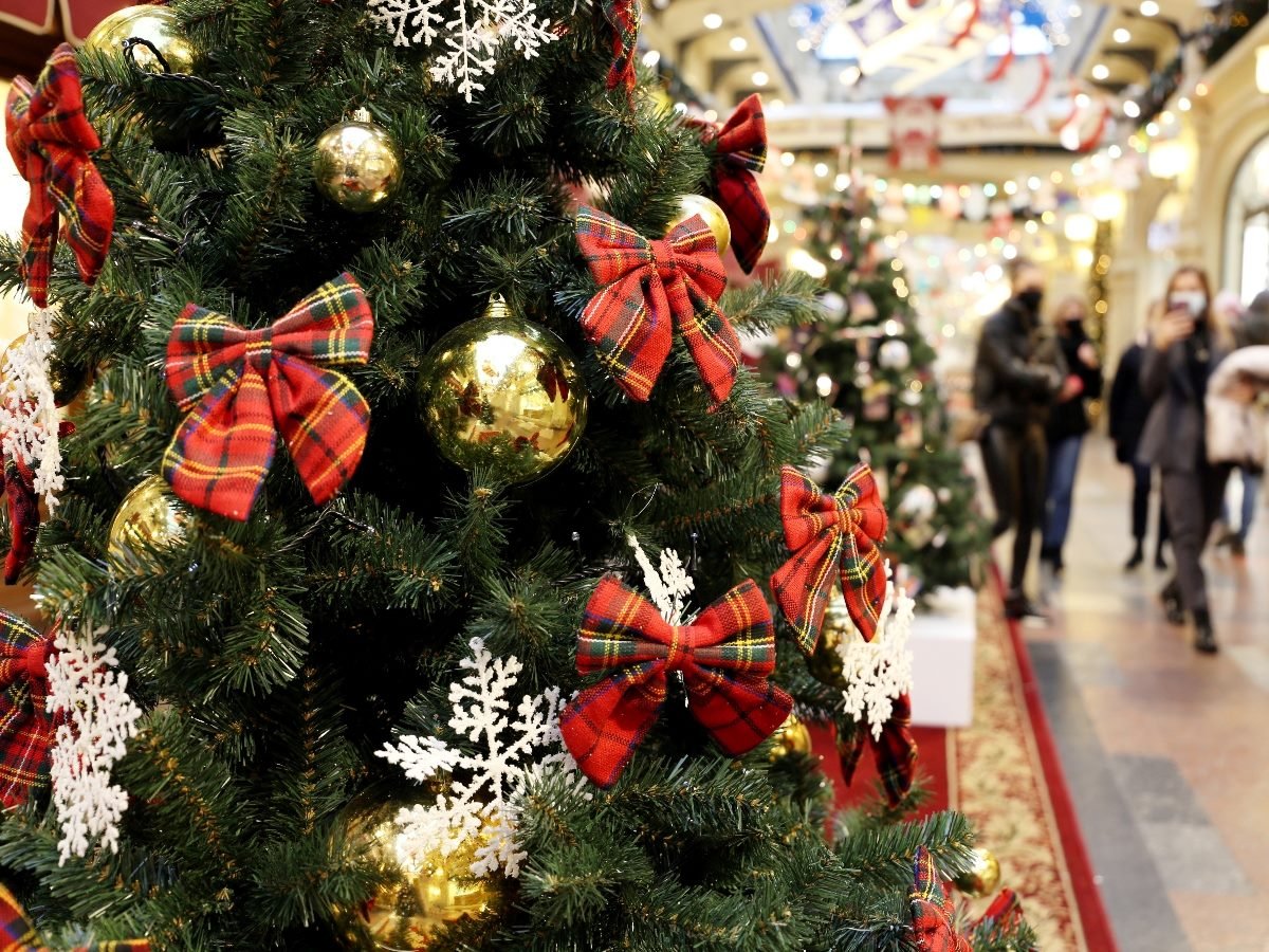 A close-up of a Christmas tree decorated with red plaid bows, gold ornaments, and white snowflake decorations inside a brightly lit shopping mall, with holiday shoppers walking in the blurred background.