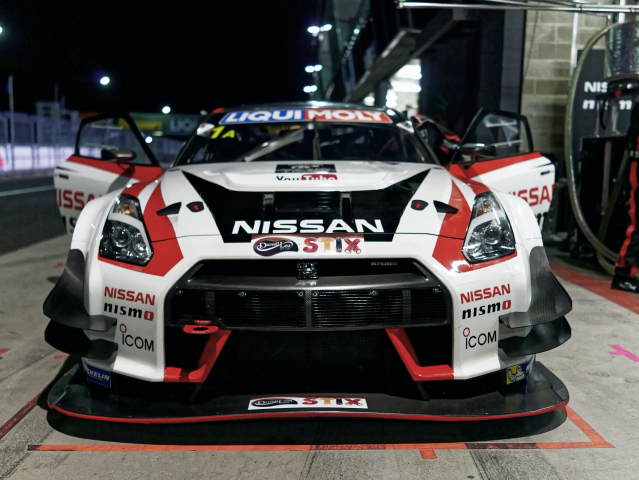 A Nissan race car in the pits. A head on view of the car.  The cars is colored white, red and black