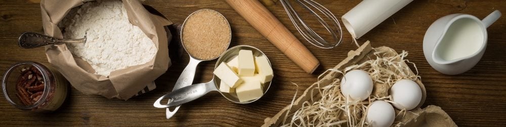 Baking ingredients, including flour in a bag, chunks of butter in a cup, sugar in a cup and three eggs in straw, a rolling pin, whisk an white pitcher are visible in the upper right.