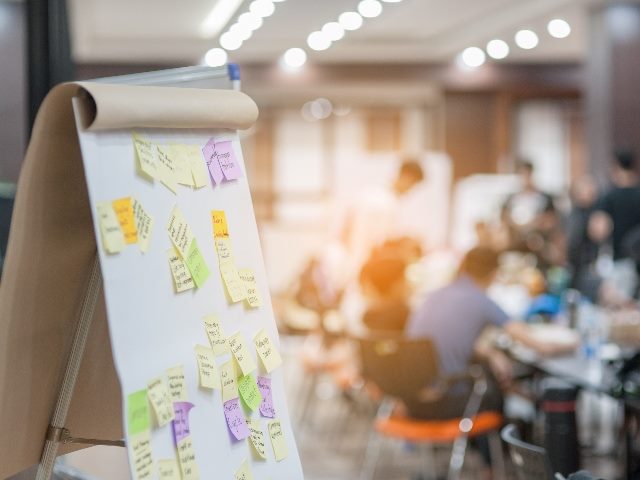 Business people in the background meeting at office and use post it notes on board to share ideas. 