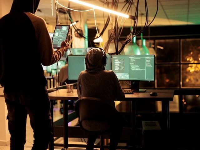 Hackers, one sitting at a computer terminal and the other holding a laptop standing behind the other hacker, running spyware code on computer screen.