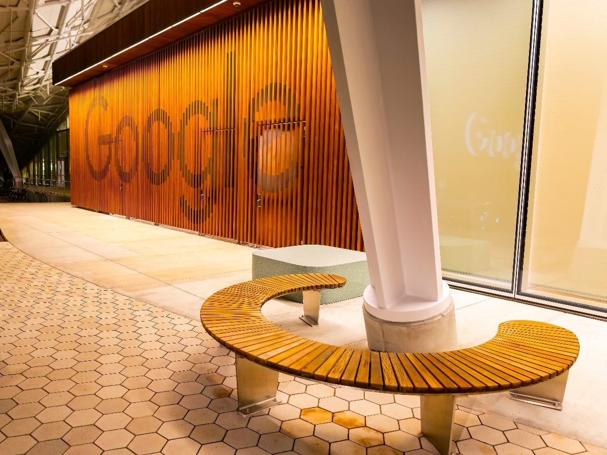 nterior view of a Google office corridor featuring a large wooden slat wall with the Google logo integrated into the design, curved wooden bench seating around a structural column, hexagonal tile flooring and warm ambient lighting in a modern architectural space.