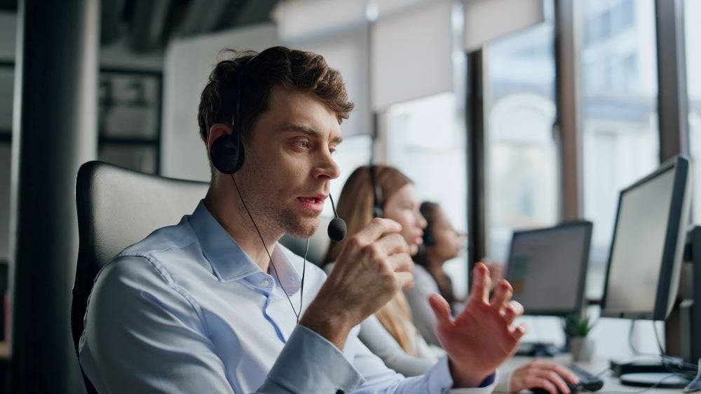A customer service agent wearing a headset and sitting in front of a computer screen in a contact center with other frontline workers talks to a customer.